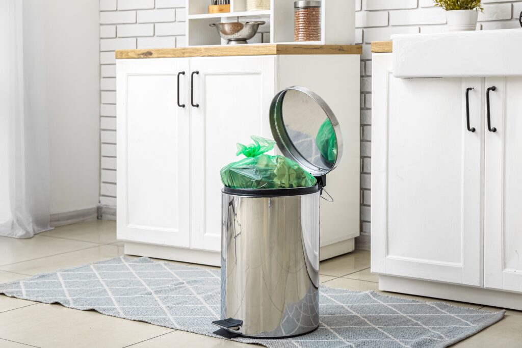 A stainless steel trash can with an open lid and a green trash bag inside is placed on a patterned grey rug in a kitchen. The kitchen features white cabinets with black handles, a wooden countertop, and a white brick wall backsplash.