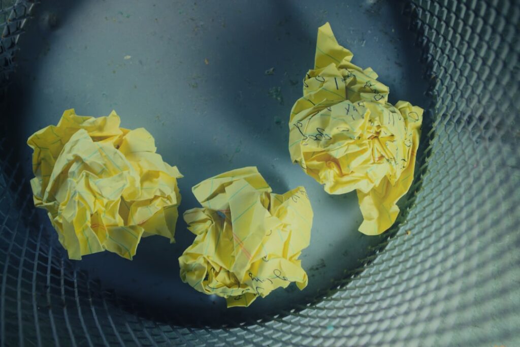 Three crumpled yellow paper balls with faint writing on them sit at the bottom of a textured, metallic wastebasket. The papers are slightly illuminated, creating a contrast with the wastebasket's darker interior.