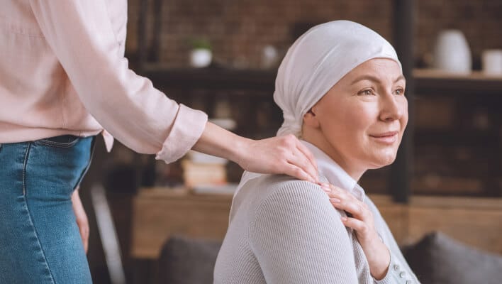 A woman wearing a headscarf sits and smiles gently while another person stands beside her, offering comfort by resting a hand on her shoulder. The scene suggests support and care.