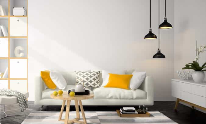 A modern living room with a white sofa, yellow and gray pillows, hexagon-patterned rug, wooden coffee table with tea set and lemons, pendant lights, open shelving, and white sideboard with a plant.