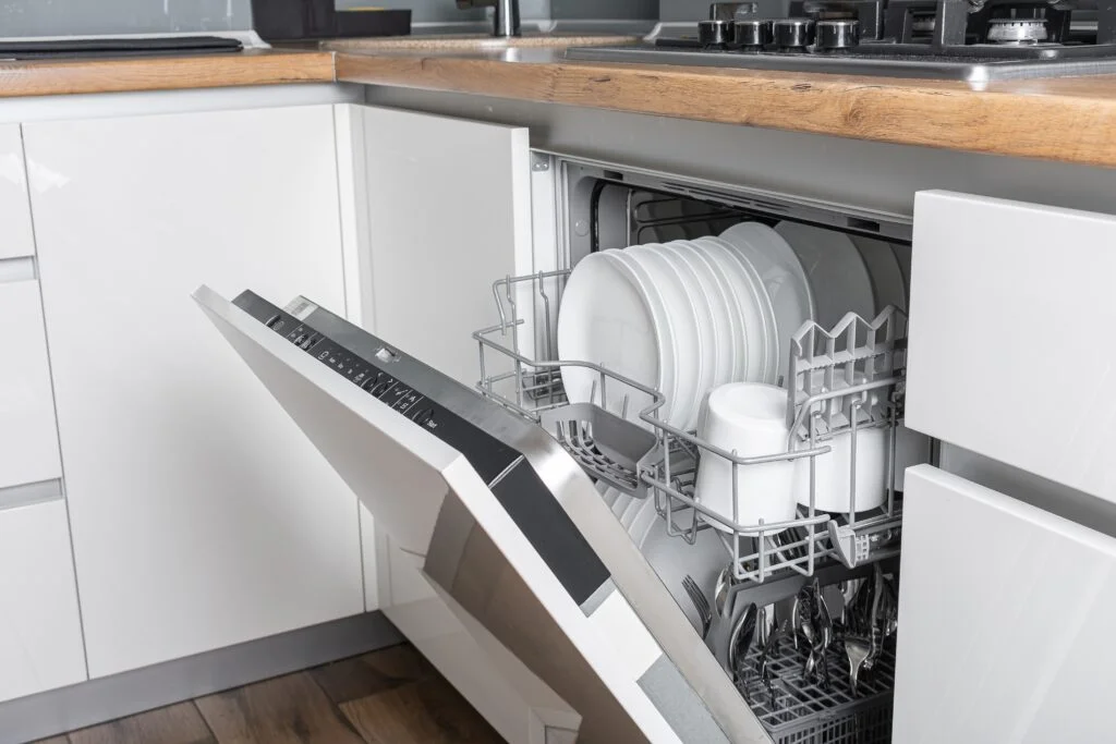 Open built-in dishwasher in a modern kitchen, loaded with clean white dishes and cups.