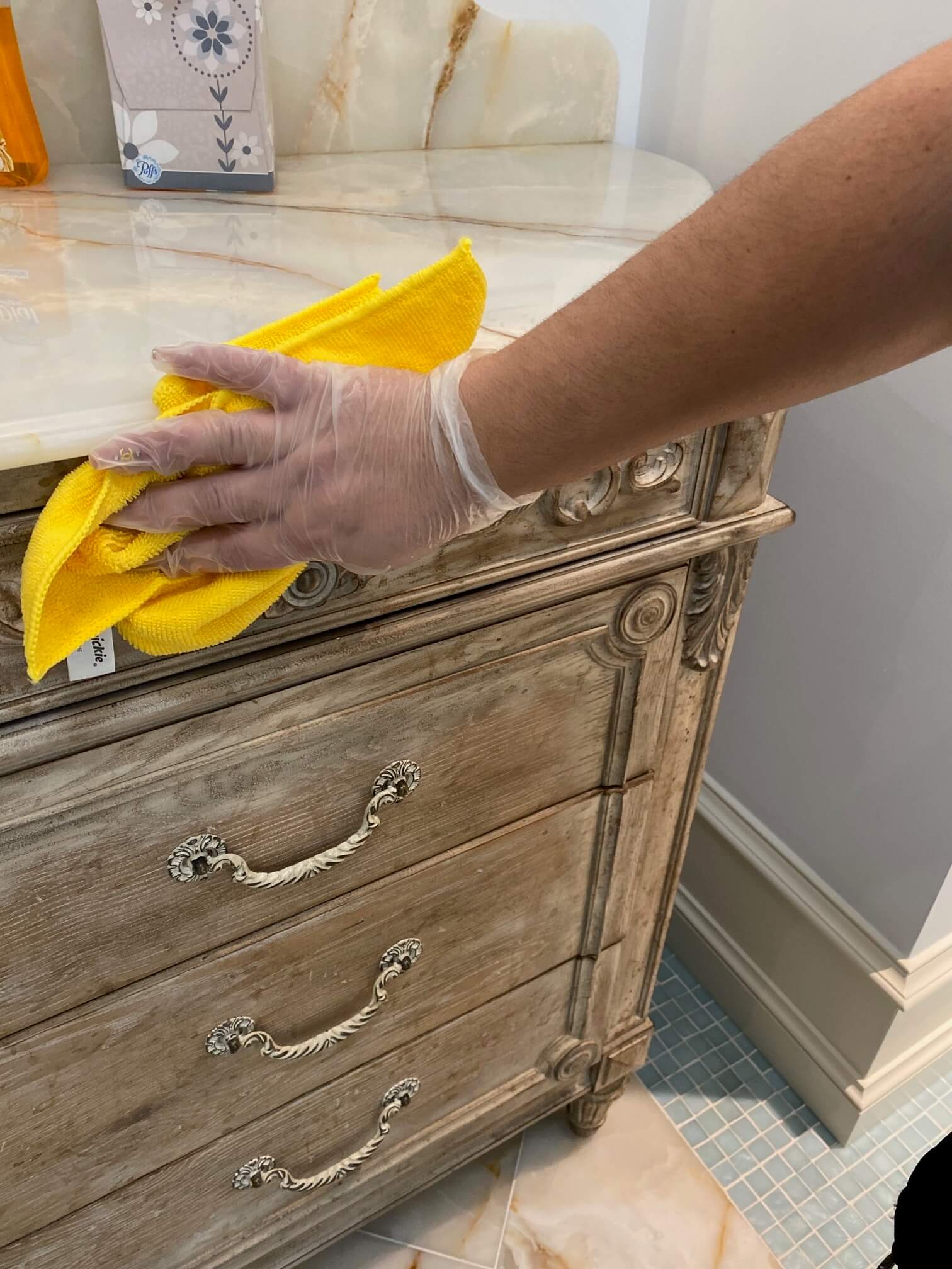 A person wearing a disposable glove wipes a wooden dresser with a yellow cloth in a bathroom, cleaning the marble countertop.