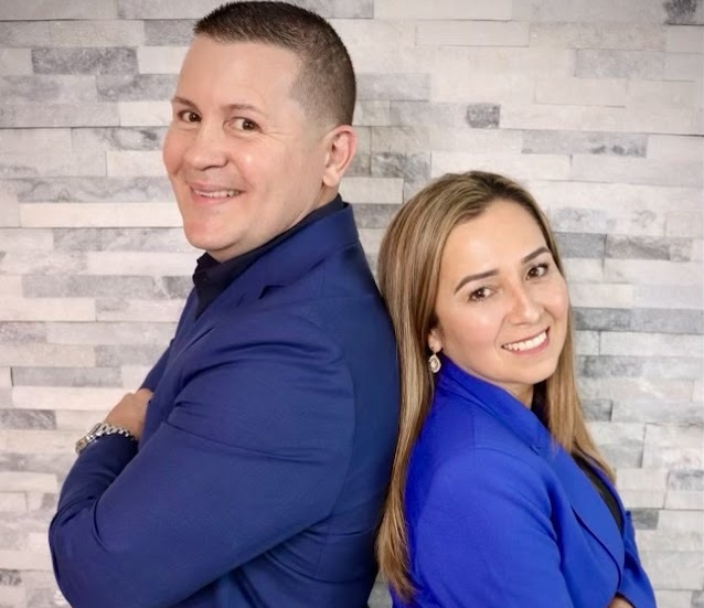 A man and a woman stand back to back, both smiling at the camera. They are dressed in blue blazers and are posed against a light gray stone tile wall.
