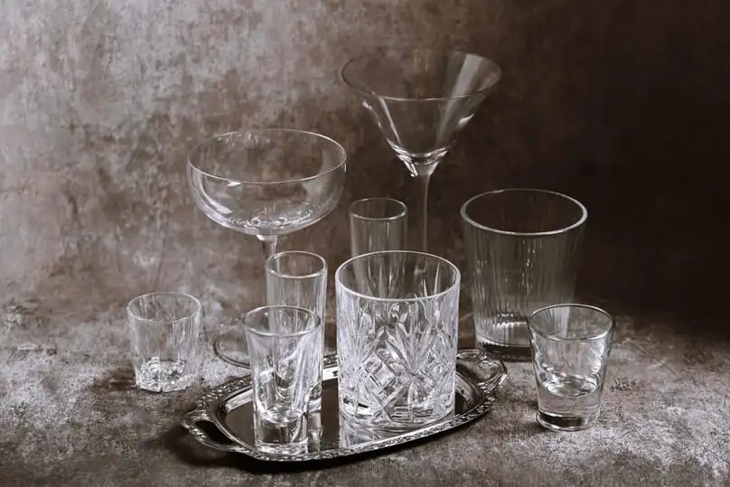 A collection of sparkling, diverse crystal glasses, including wine glasses, martini glasses, and tumblers, are arranged on a silver tray against a textured background, highlighting the need to know how to clean crystal glasses.