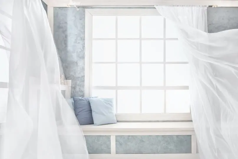 Long white sheer curtains gently billow in a breeze beside a large window with a cushioned seat below, suggesting the importance of knowing how to wash curtains to keep them fresh.