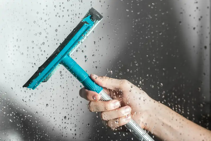 A person's hand demonstrates shower squeegee tips on a wet glass surface, cleaning water droplets.