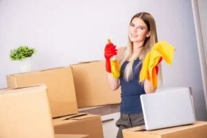 A young woman with cleaning supplies and a laptop among moving boxes, showing what is professional move out cleaning.