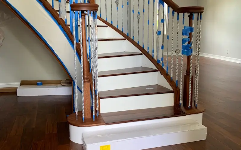 A curved staircase with dark wood steps and handrails is under renovation in a Cloud Lake home. Blue painter’s tape covers parts of the metal balusters and wood to protect them, while the floor and nearby surfaces are also taped for safety.