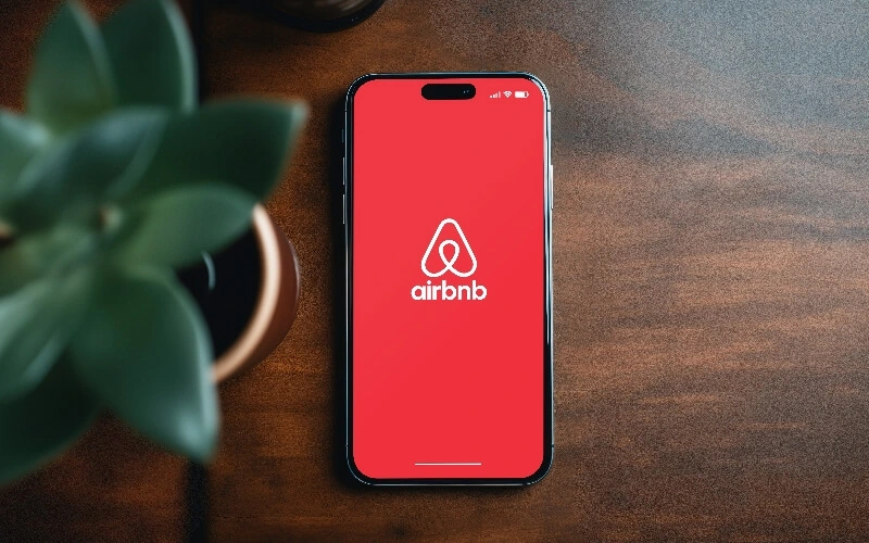 A smartphone displaying the red Airbnb app launch screen with the Airbnb logo, placed on a wooden surface next to a green potted plant, evokes a sense of adventure—like planning your next getaway along the Gulf Stream.