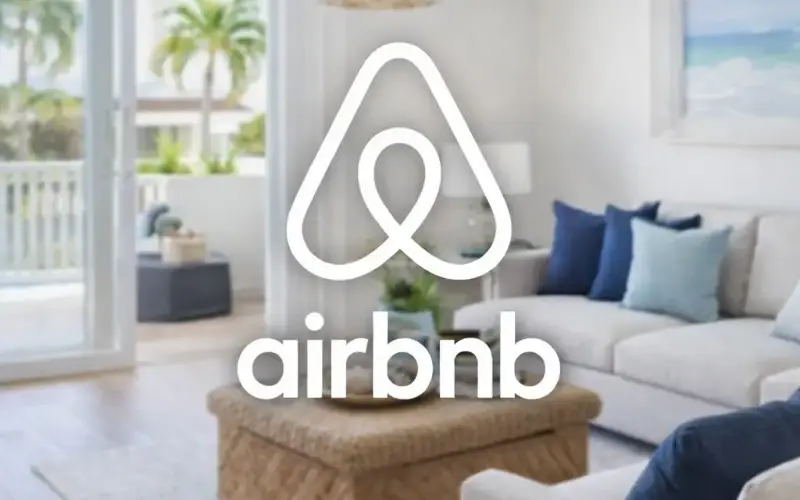 The Airbnb logo is centered over a bright, modern living room in Royal Palm Estates with a white sofa, blue pillows, a wicker table, and large windows overlooking palm trees outside.